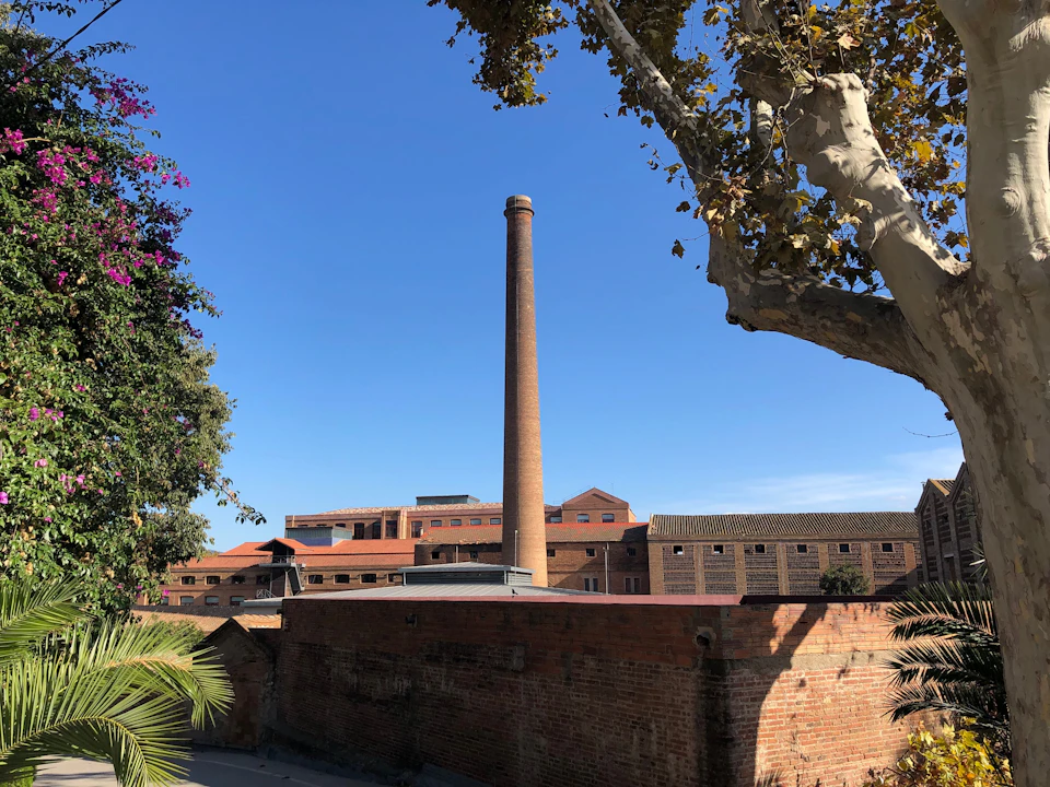 Tall brick chimney amidst red brick buildings, surrounded by trees and clear blue sky.