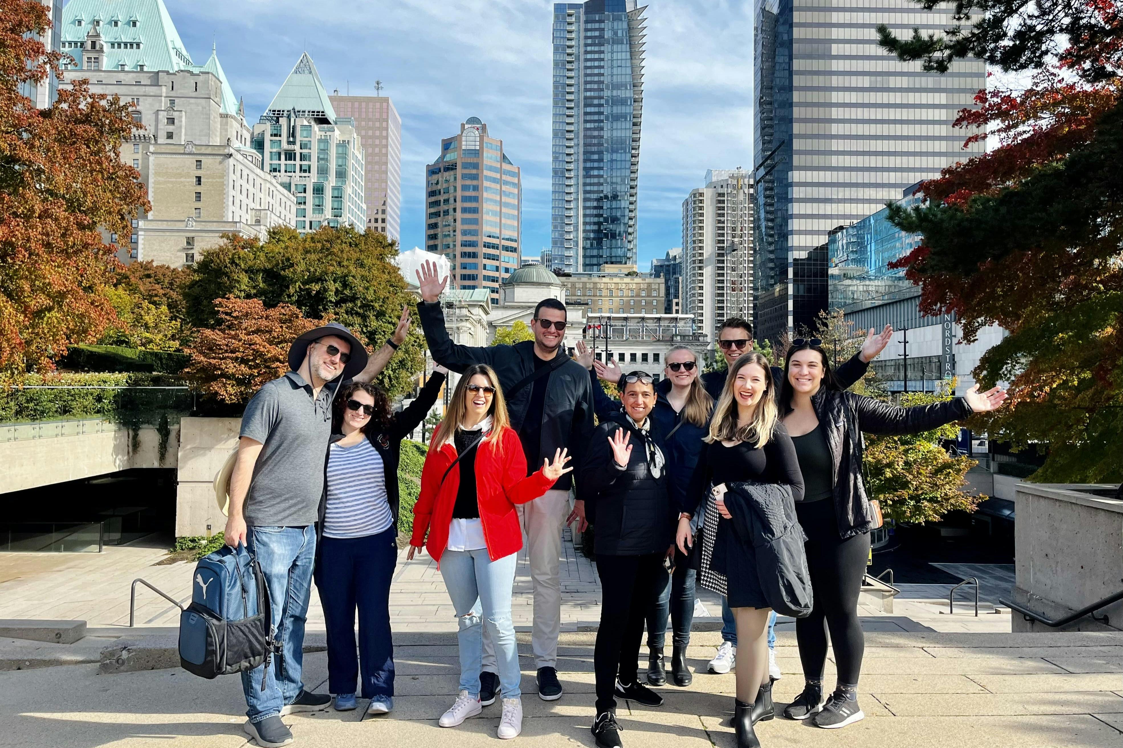 Skupina lidí pózujících a usmívajících se venku v panoramatu města s vysokými budovami, stromy a jasně modrou oblohou v pozadí.