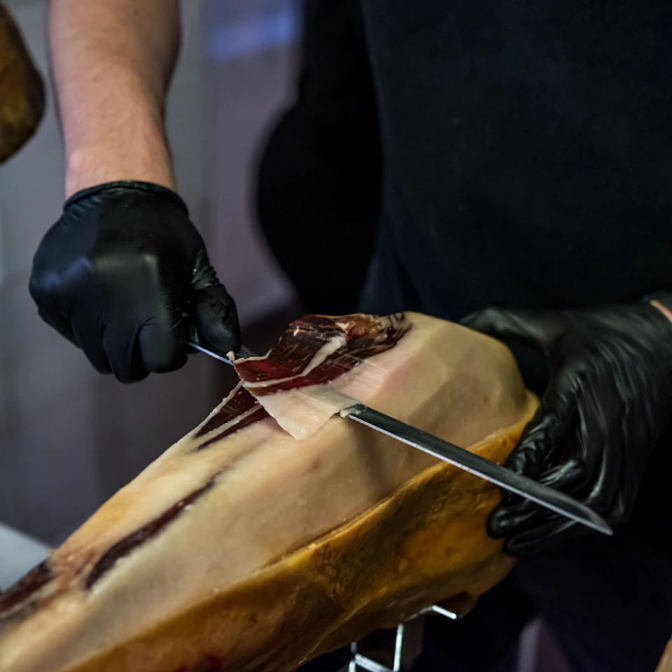 A person wearing black gloves slicing a piece of cured meat from a ham leg using a knife.