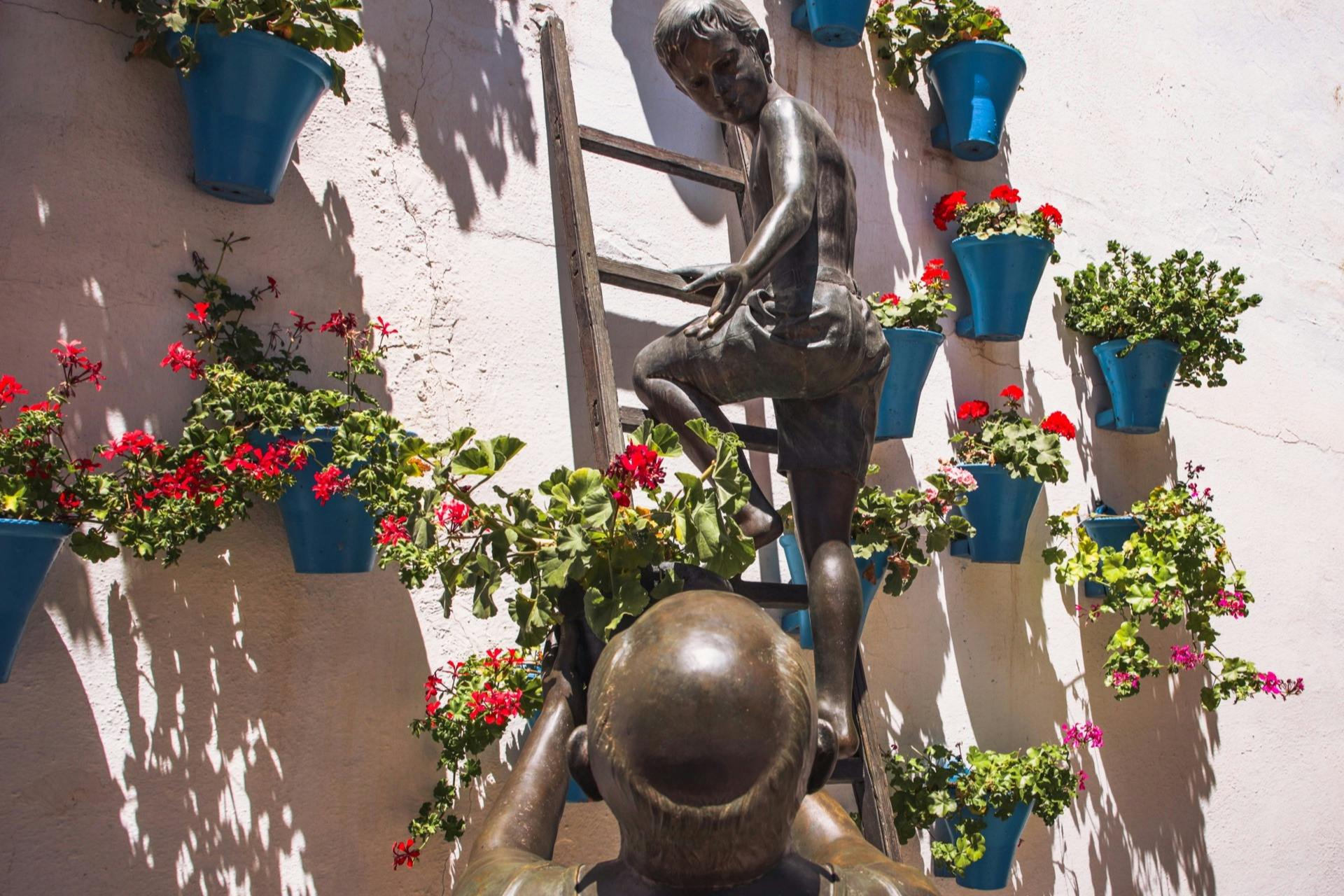 Bronze statues of a child on a ladder and an adult supporting them, surrounded by blue flower pots with red and pink blossoms.