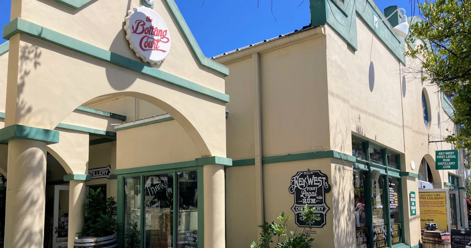 A light beige building with green trim featuring signs for 'The Bottling Court' and 'Key West Legal Rum Distillery.'