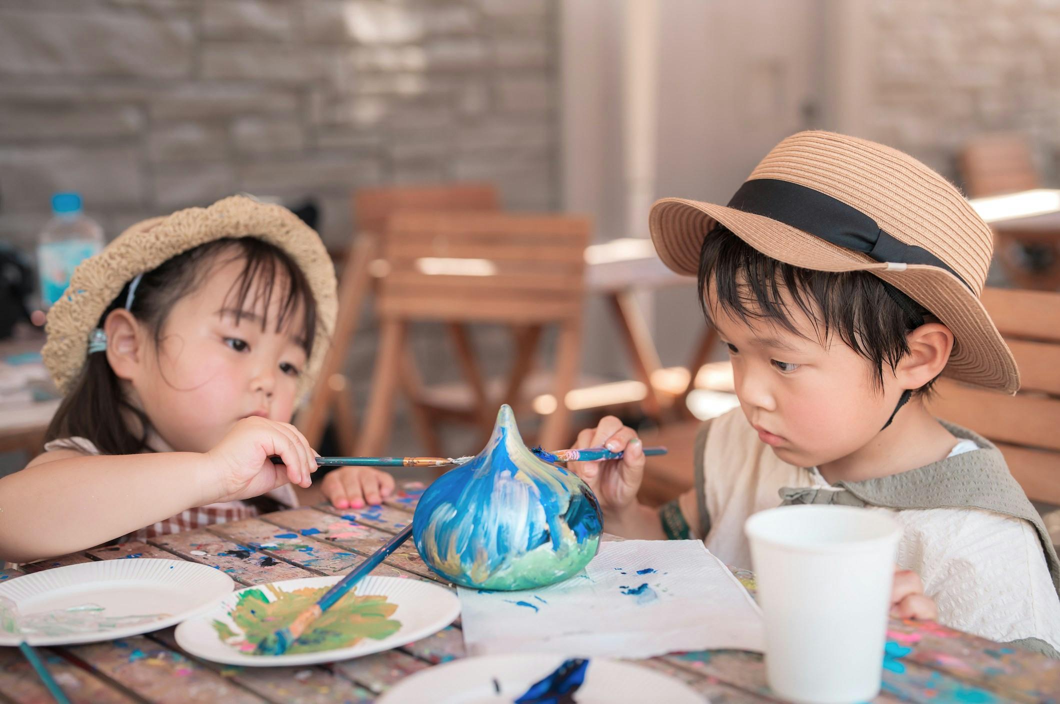 A child wearing a straw hat paints a gourd with blue and green colors at a table outdoors. Art supplies are visible.