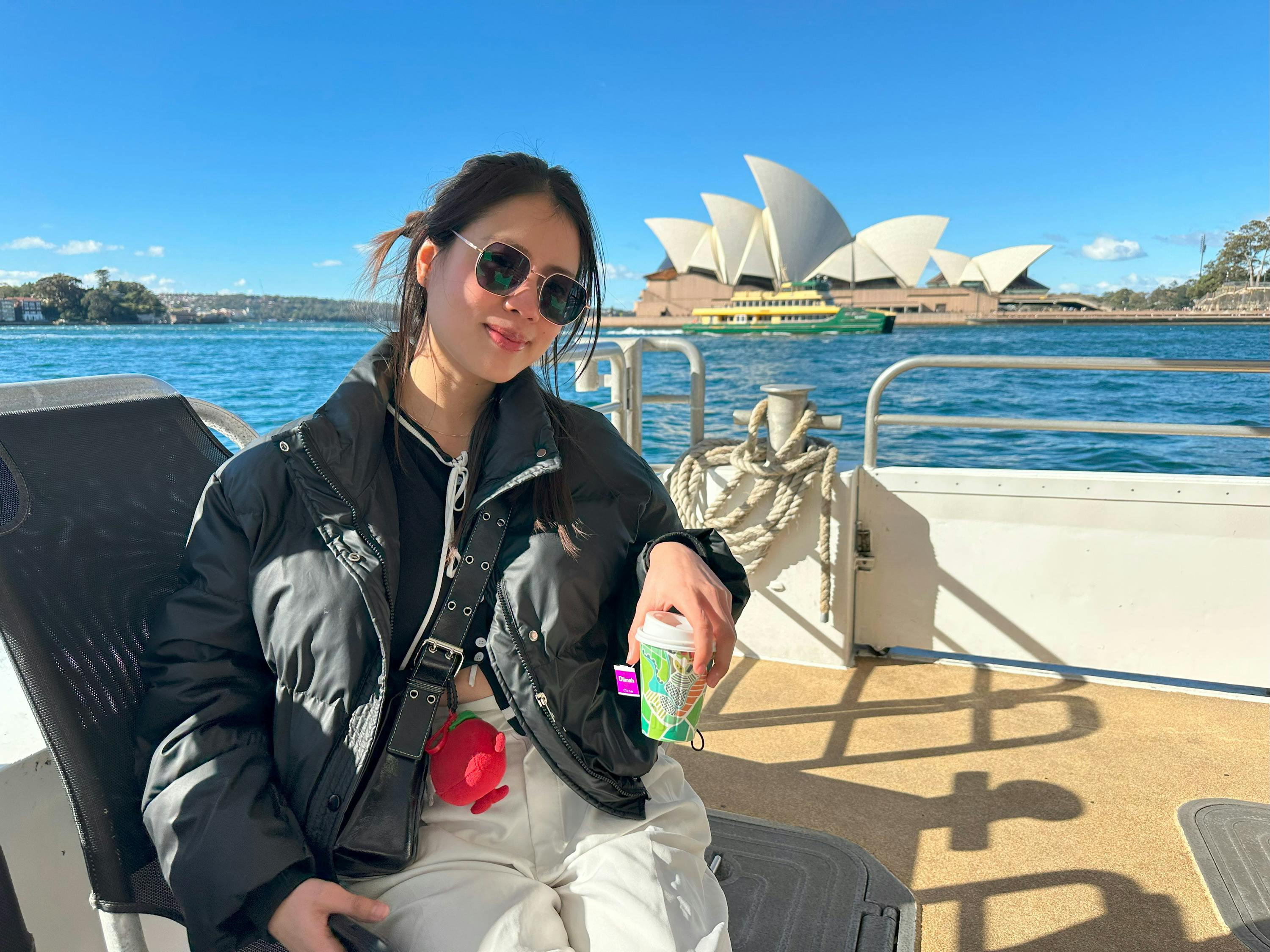 A person wearing sunglasses and a black jacket holding a cup on a boat with the Sydney Opera House in the background.