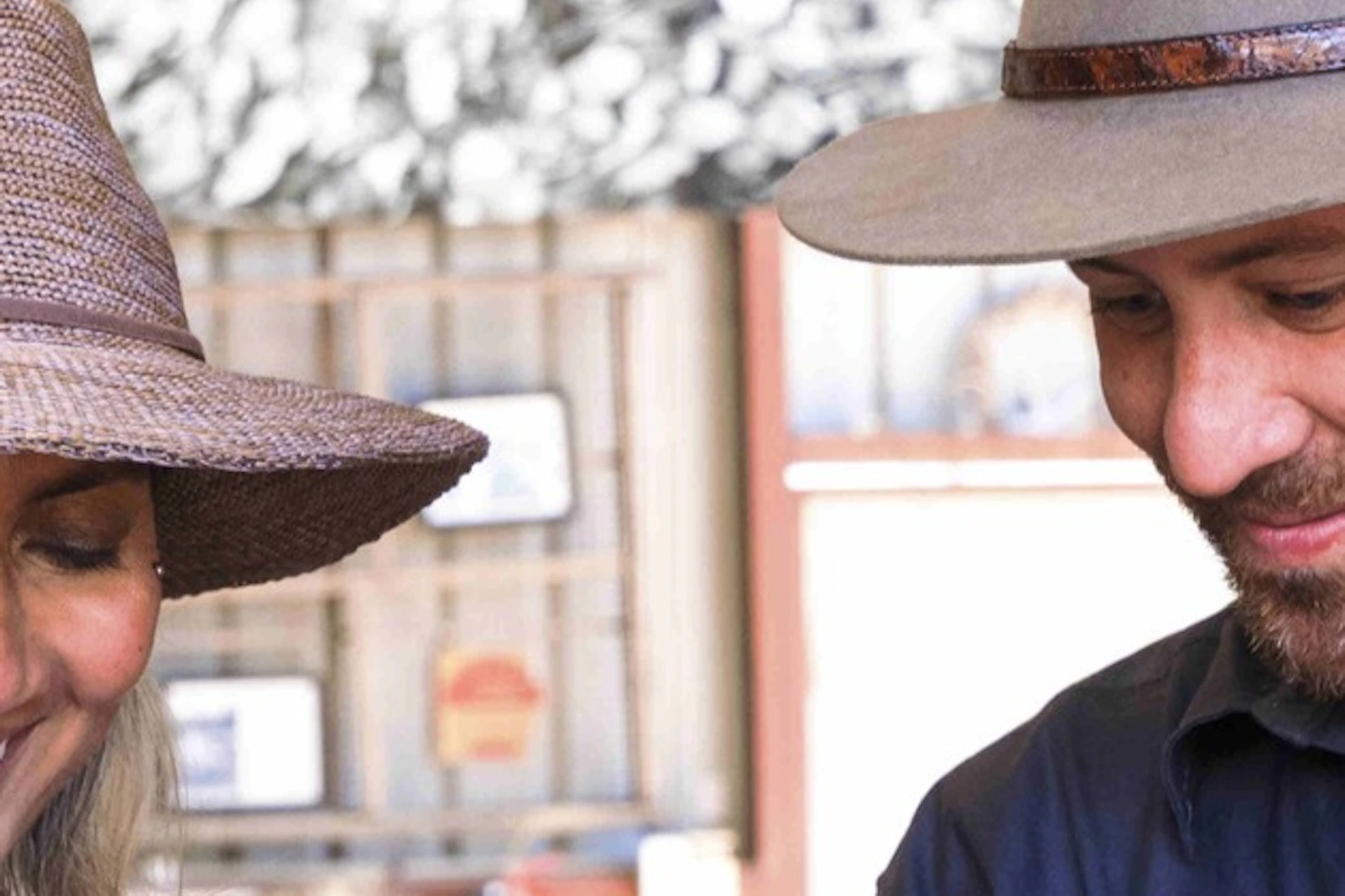 Two individuals wearing hats interact while preparing oysters at a rustic setting with oysters and tools on a table.