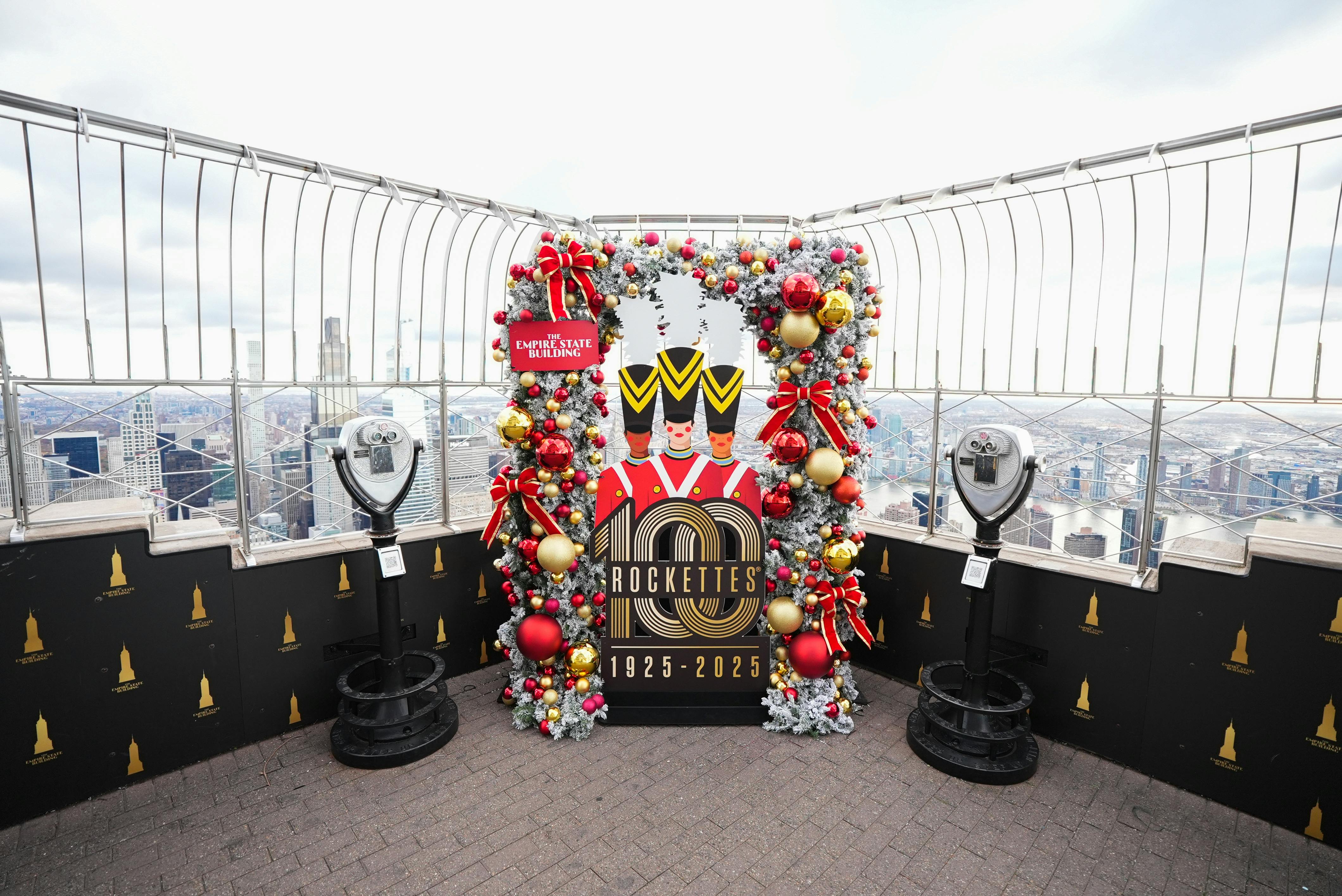 A festive display on an observation deck with a Rockettes 100th anniversary sign surrounded by colorful decorations and binocular viewers.