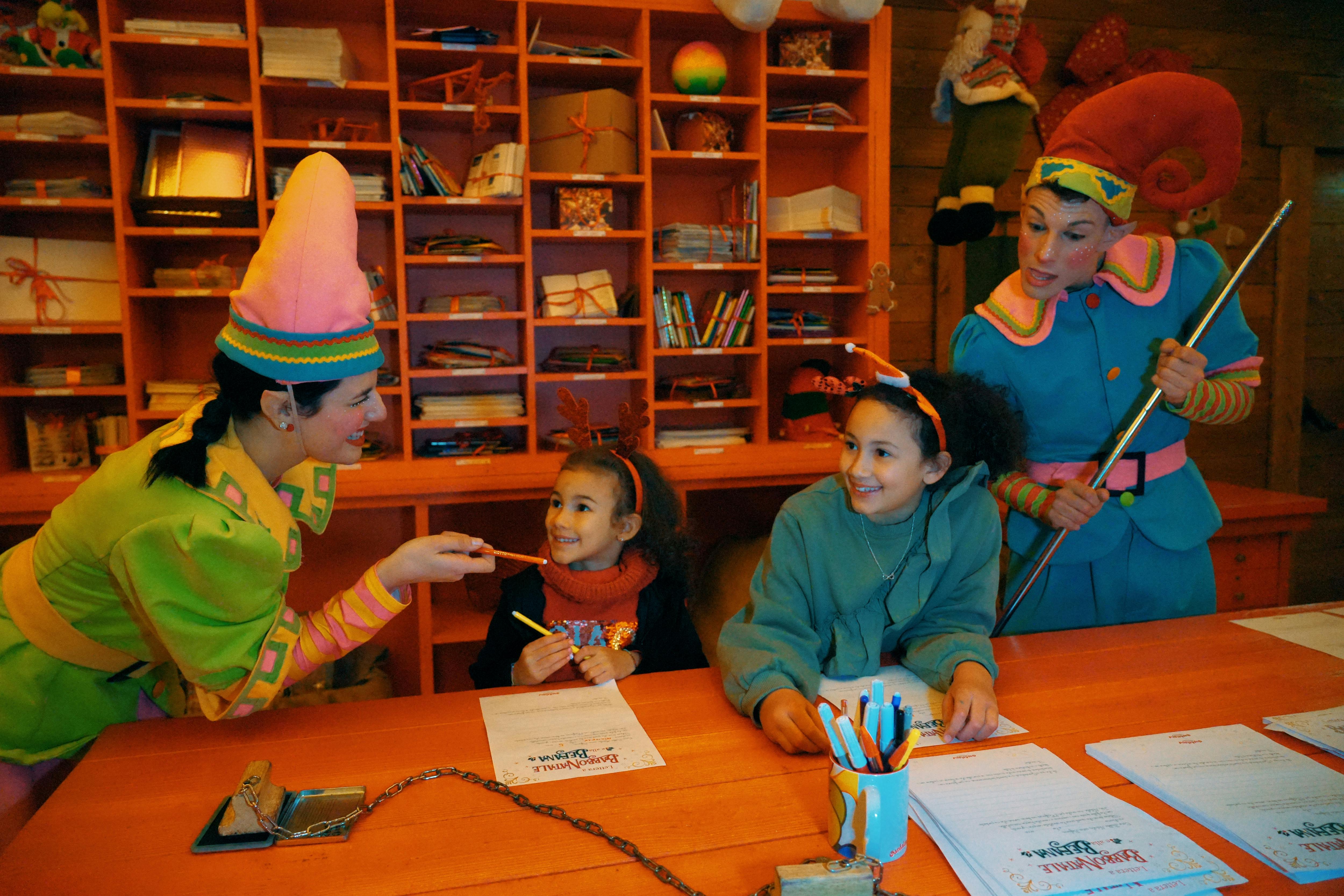 Dvě děti u pracovního stolu se usmívají na dva dospělé, oblečené jako elfové, v barevné slavnostní místnosti s uspořádanými policemi.