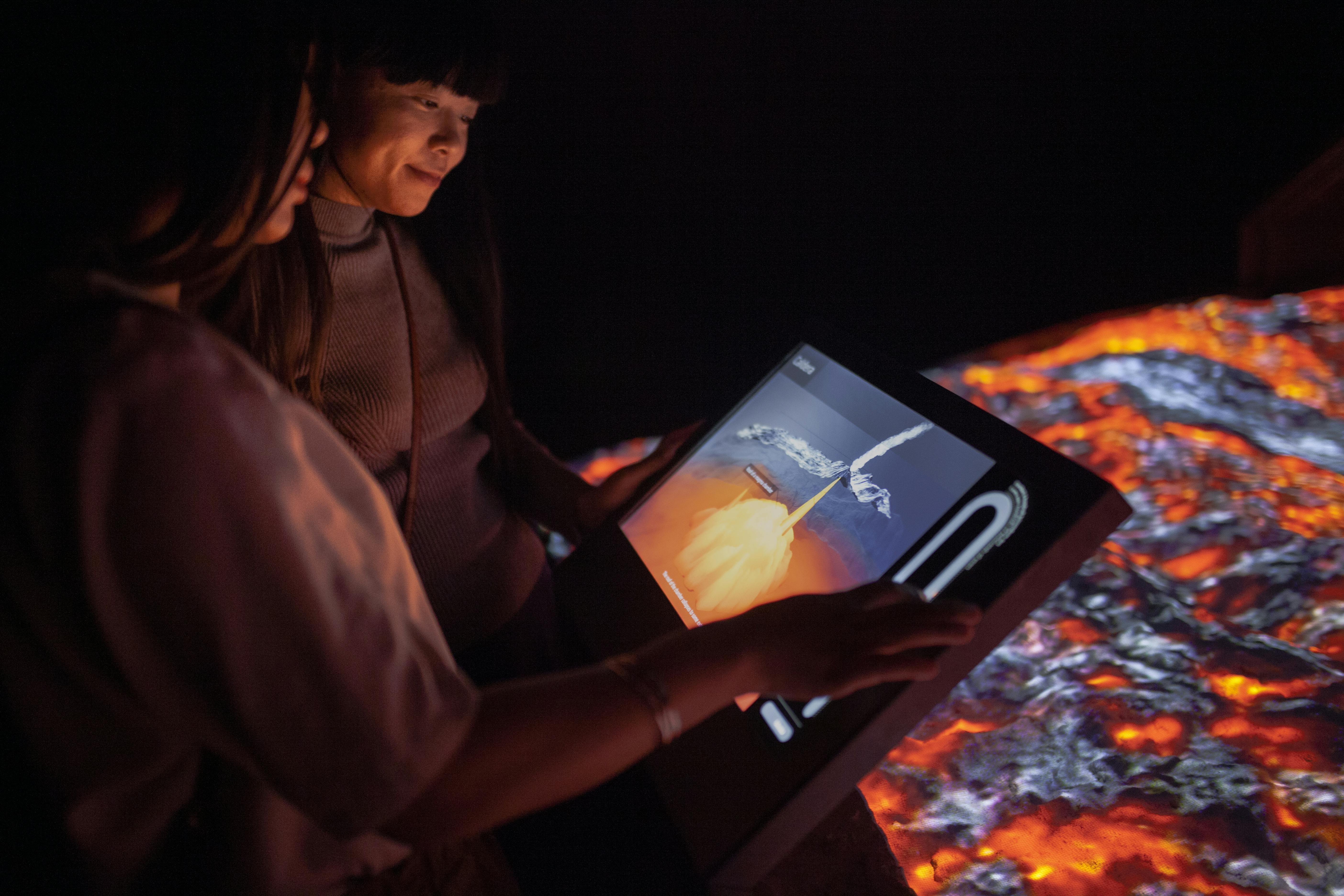 두 사람이 분출하는 화산의 이미지를 보여주는 어두운 환경에서 터치스크린 디스플레이와 상호 작용합니다.