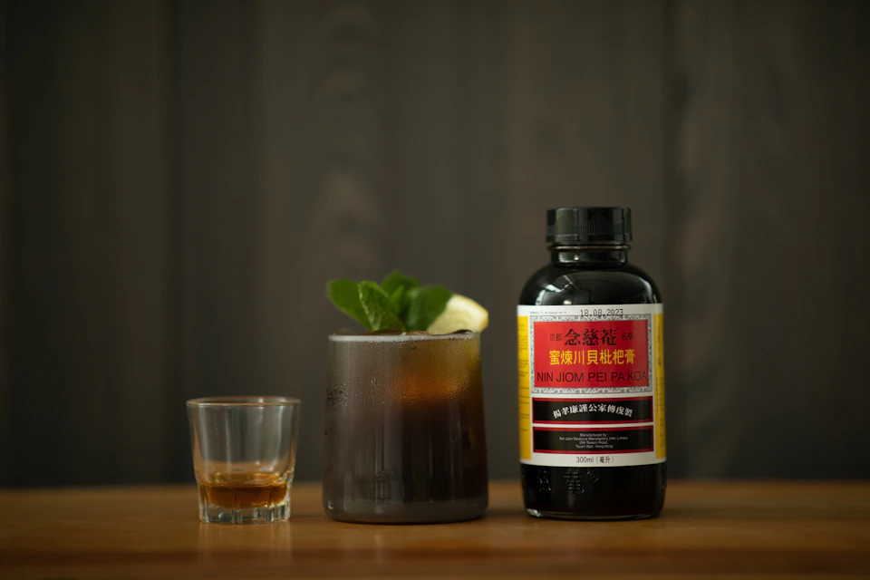 A bottle of Nin Jiom Pei Pa Koa next to a glass with a dark beverage, mint leaves, and a lime wedge, and a shot glass.