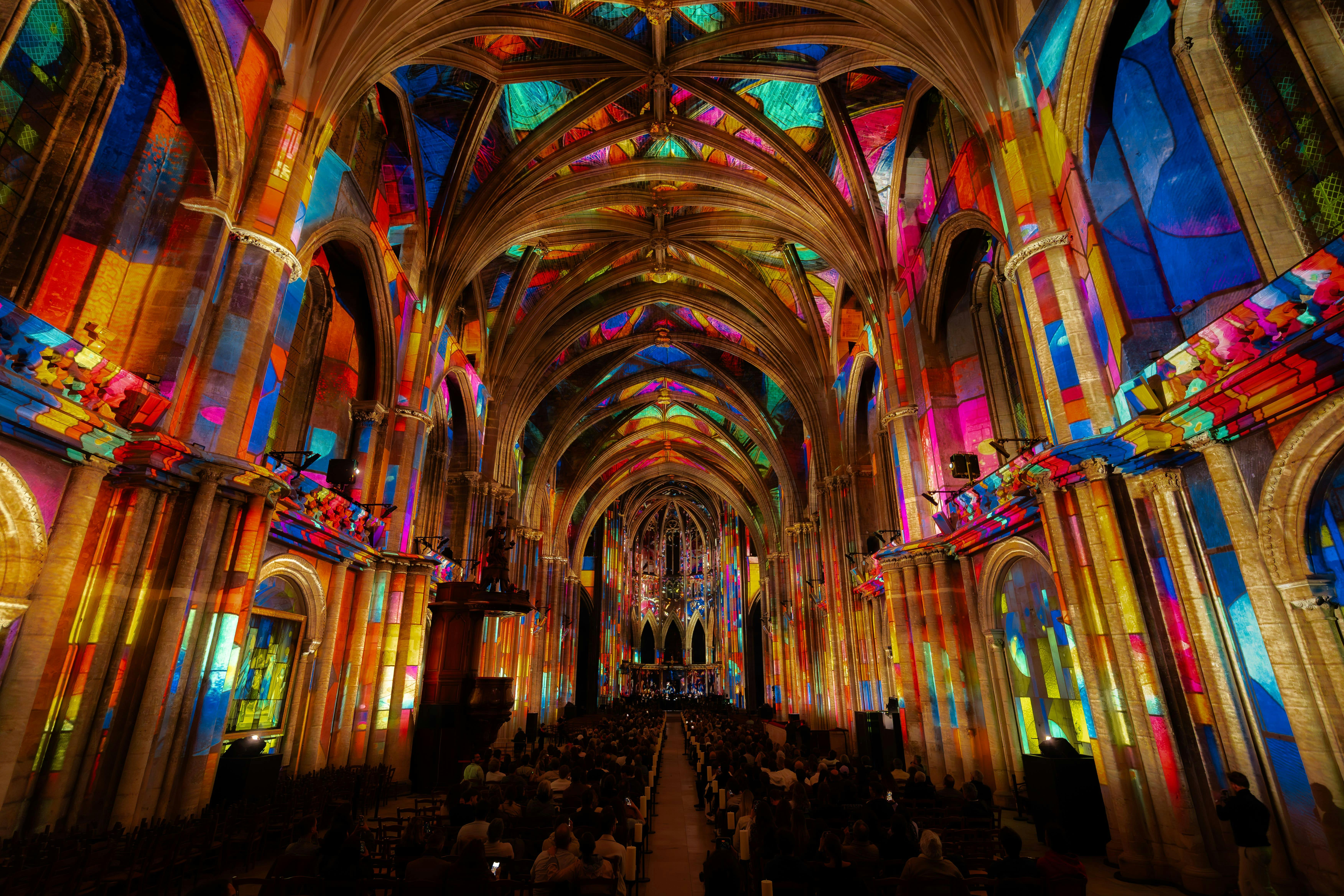 A cathedral interior with arched ceilings illuminated by vibrant, multicolored lights, and people seated along the aisle.