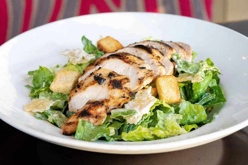 Grilled chicken breast slices on a bed of Caesar salad with croutons in a white bowl.