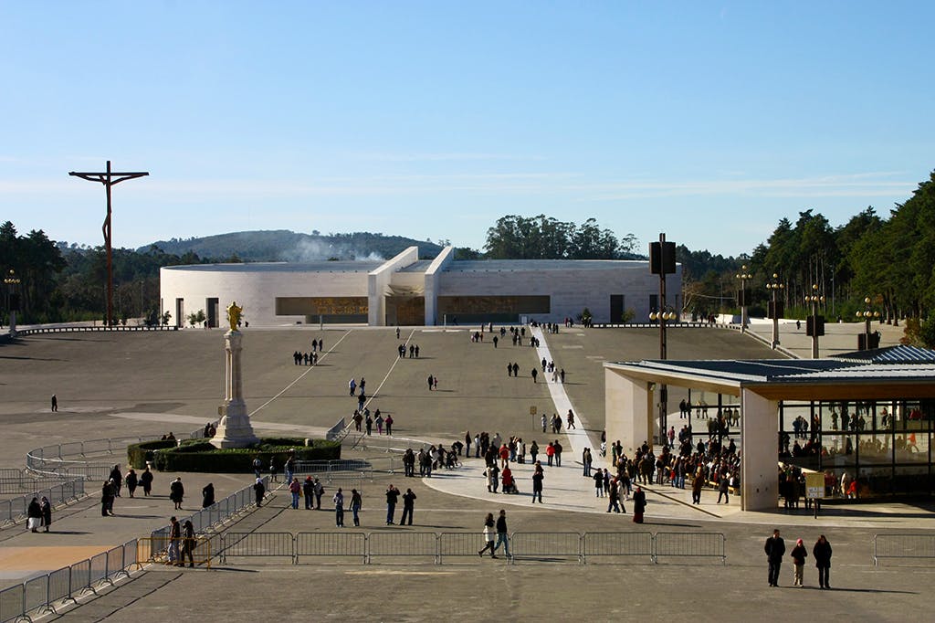 来場者が散在する広々としたオープンプラザ。 背景、彫像、前景の障壁が目立つモダンな建物。空は晴れています。