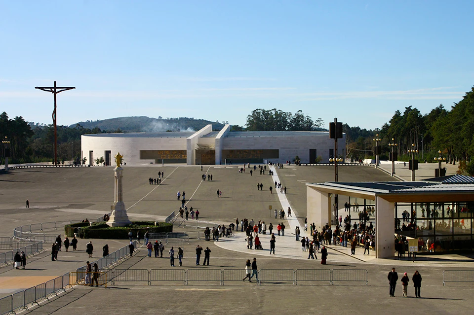 Large open plaza with scattered visitors. Prominent modern building in background, statues, and barriers in the foreground. Clear sky.