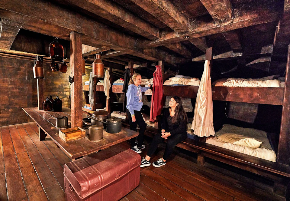 Two people inside a wooden bunkhouse with multiple bunks, bedding, and various items like pots and a suitcase around.