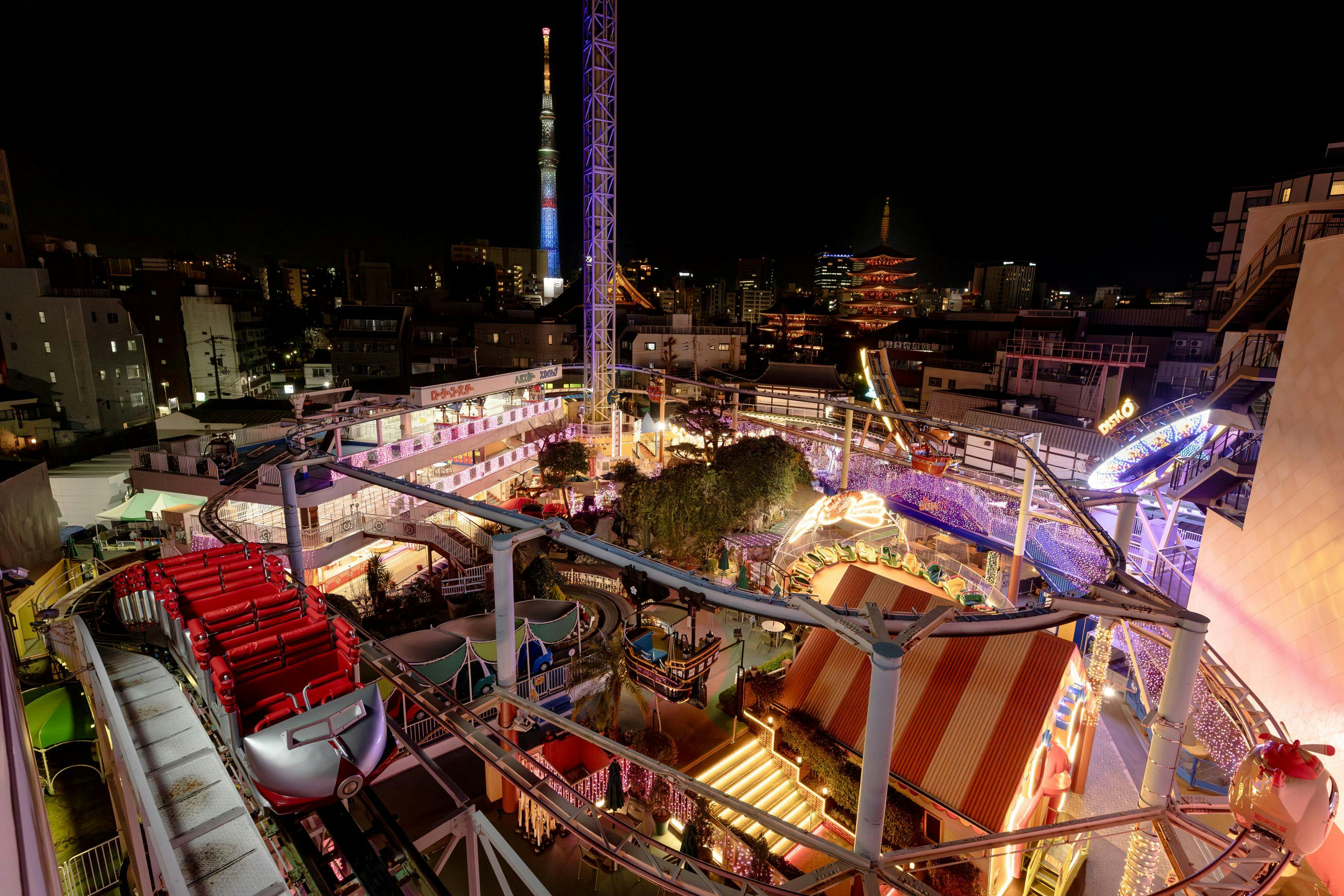 A brightly lit amusement park at night with a roller coaster, colorful decorations, and a cityscape in the background.