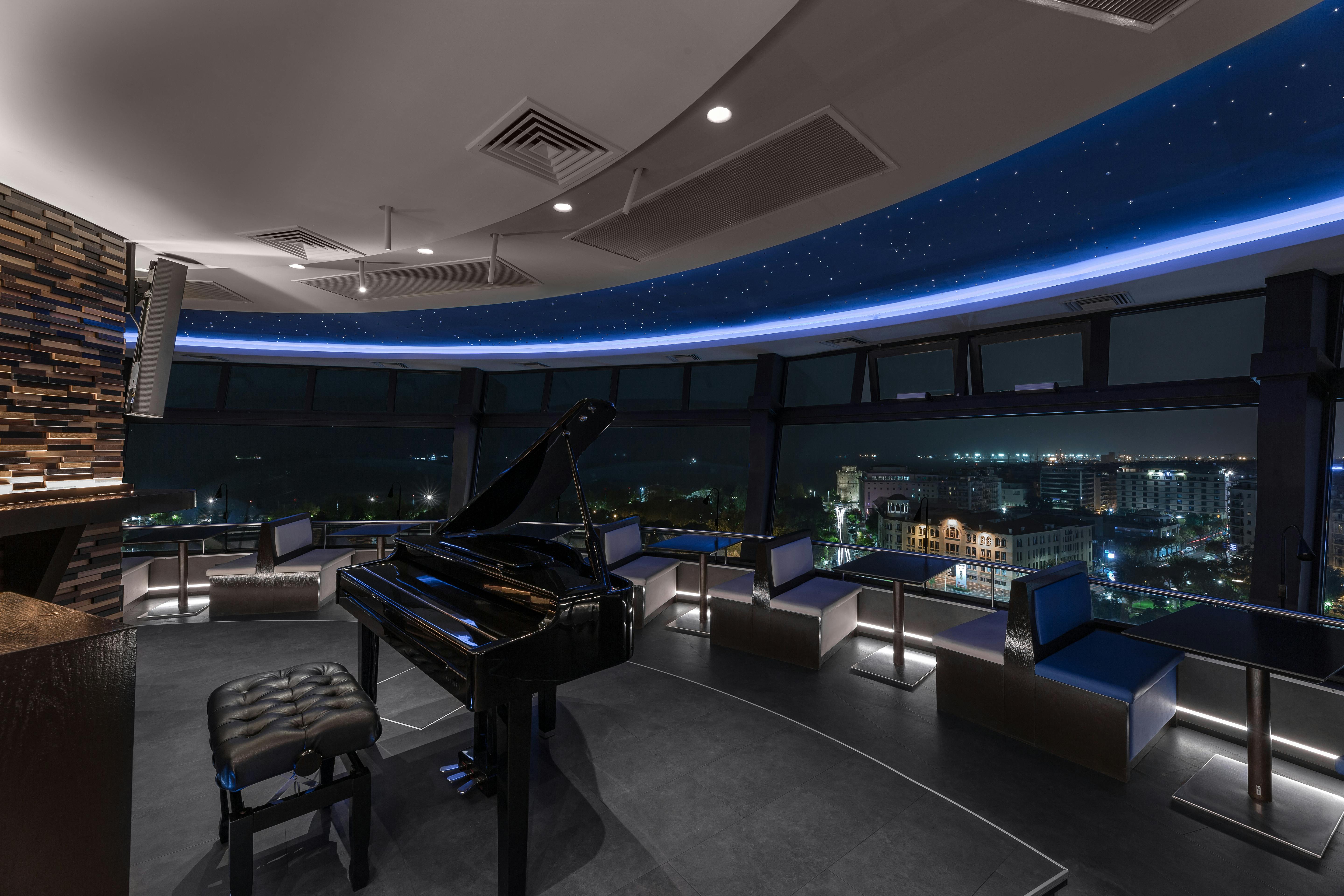 Ένα μοντέρνο δωμάτιο με μαύρο πιάνο, καθιστικό και μεγάλα παράθυρα που προσφέρουν νυχτερινή θέα σε ένα αστικό τοπίο.