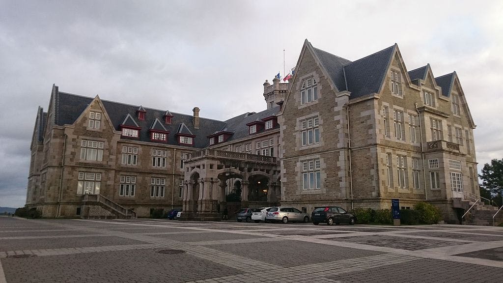 Gran edificio de piedra con muchos tejados a dos aguas y ventanas, coches aparcados delante y cielo nublado encima.