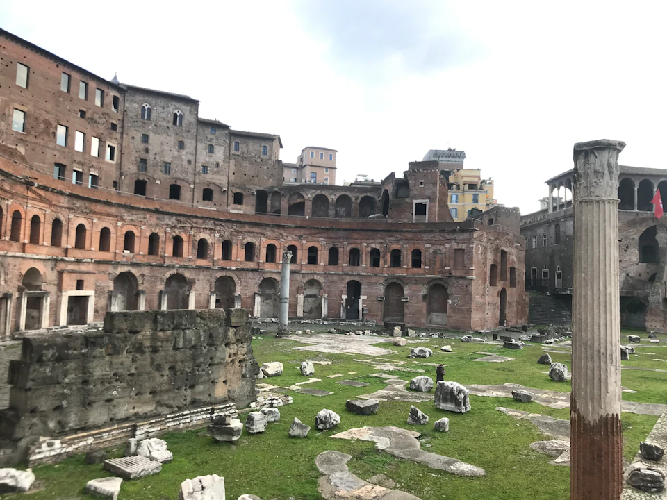 Ancient Roman ruins with arched brick structures and scattered stones on grassy ground under a cloudy sky.