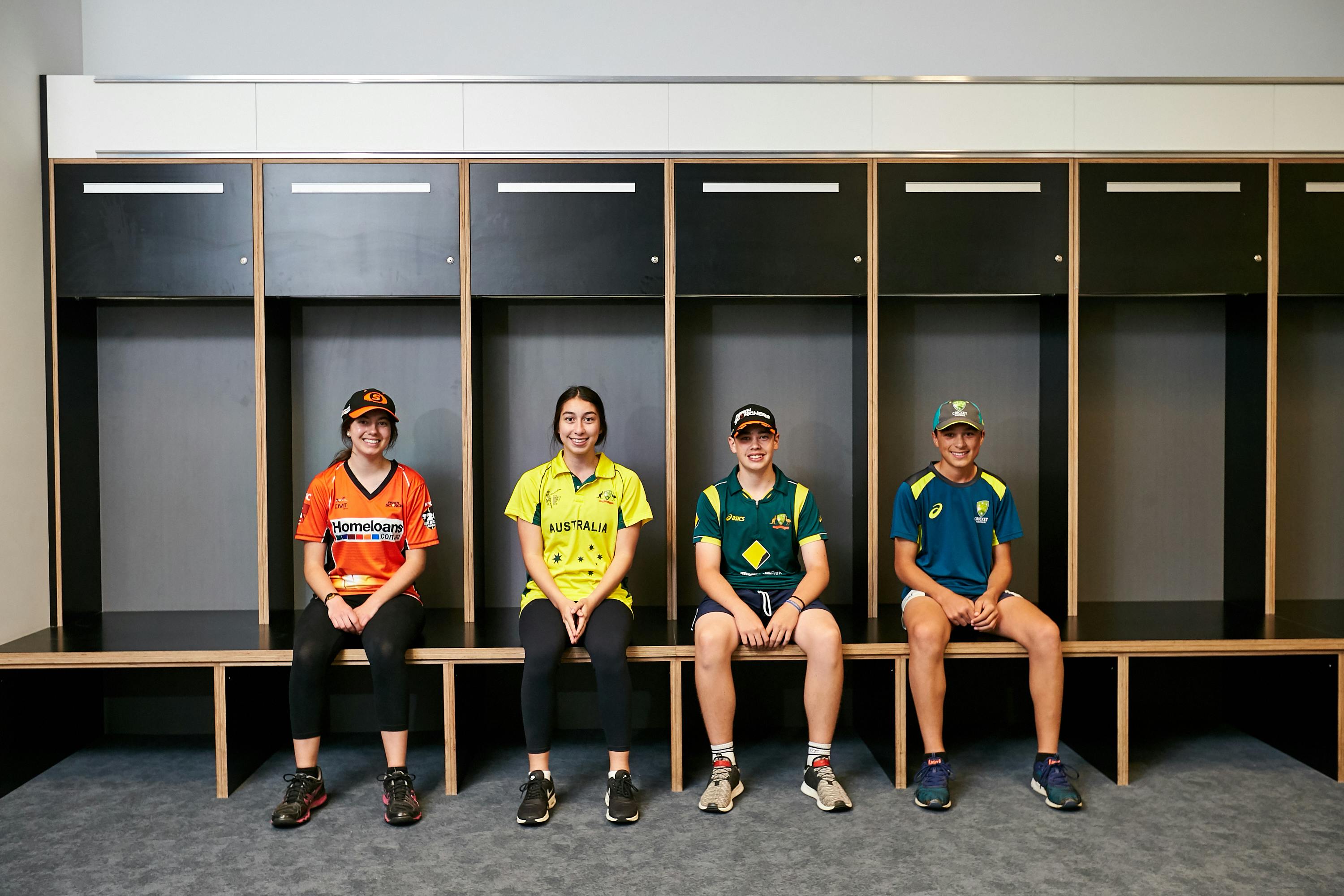 Τέσσερις άνθρωποι χαμογελούν και κάθονται σε έναν πάγκο μέσα σε ένα αποδυτήριο, φορώντας αθλητικές φανέλες και καπέλα διαφορετικού χρώματος.