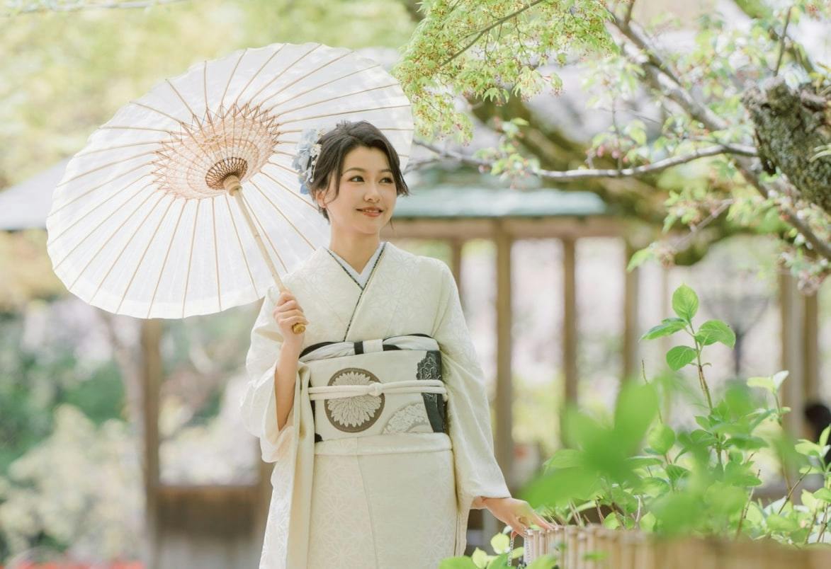 A woman wearing a white kimono holds a parasol and stands outdoors, surrounded by trees and plants.
