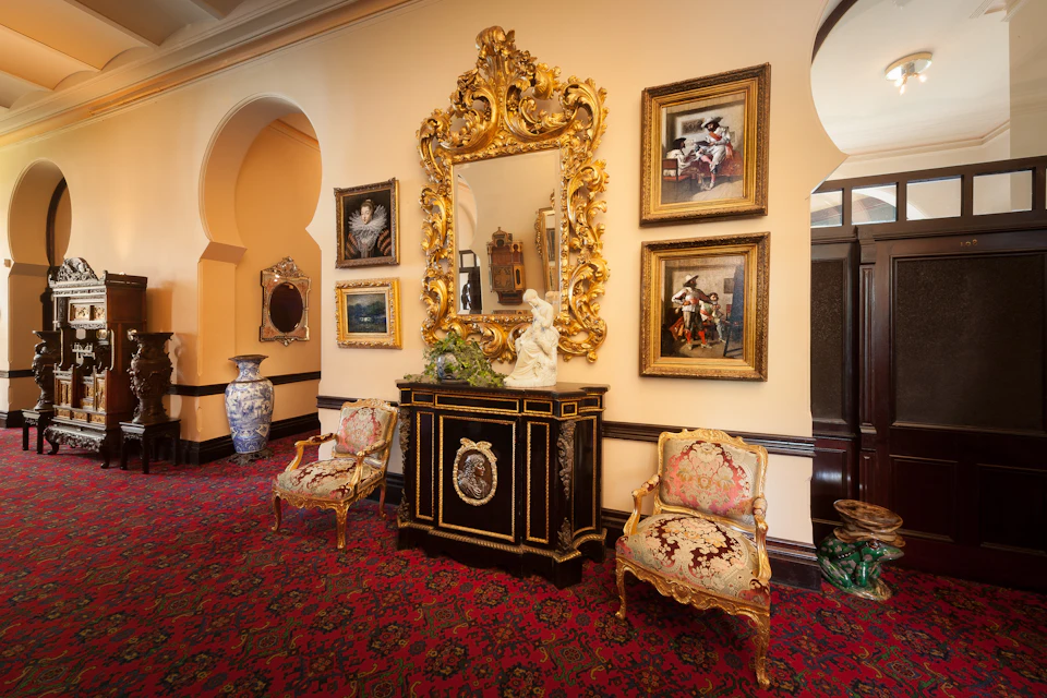 Ornate room with gold-framed mirror, framed paintings, velvet upholstered chairs, a black cabinet, and decorative vases on red carpet.