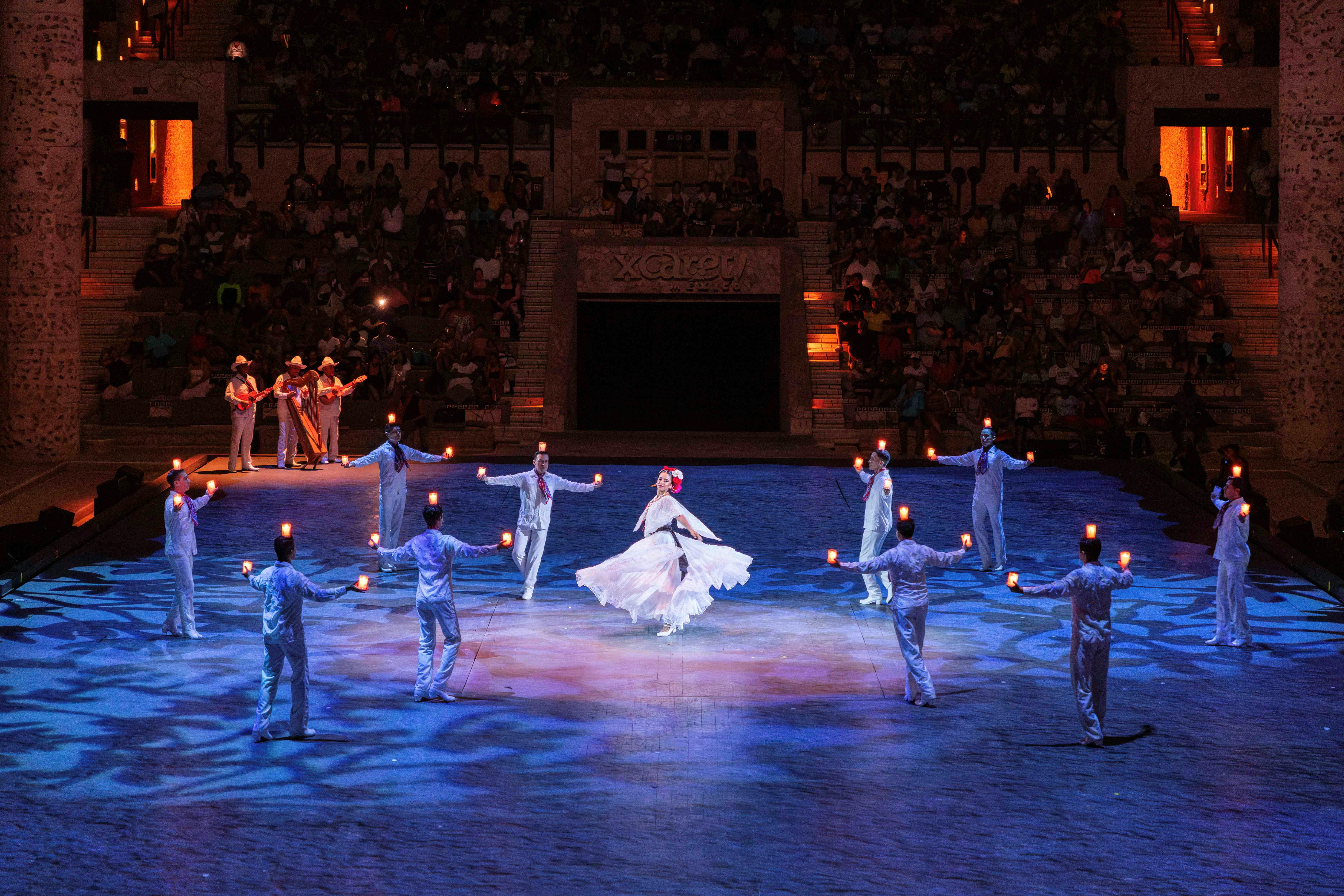Performers in traditional attire dance in a well-lit arena with a large audience watching in the background.
