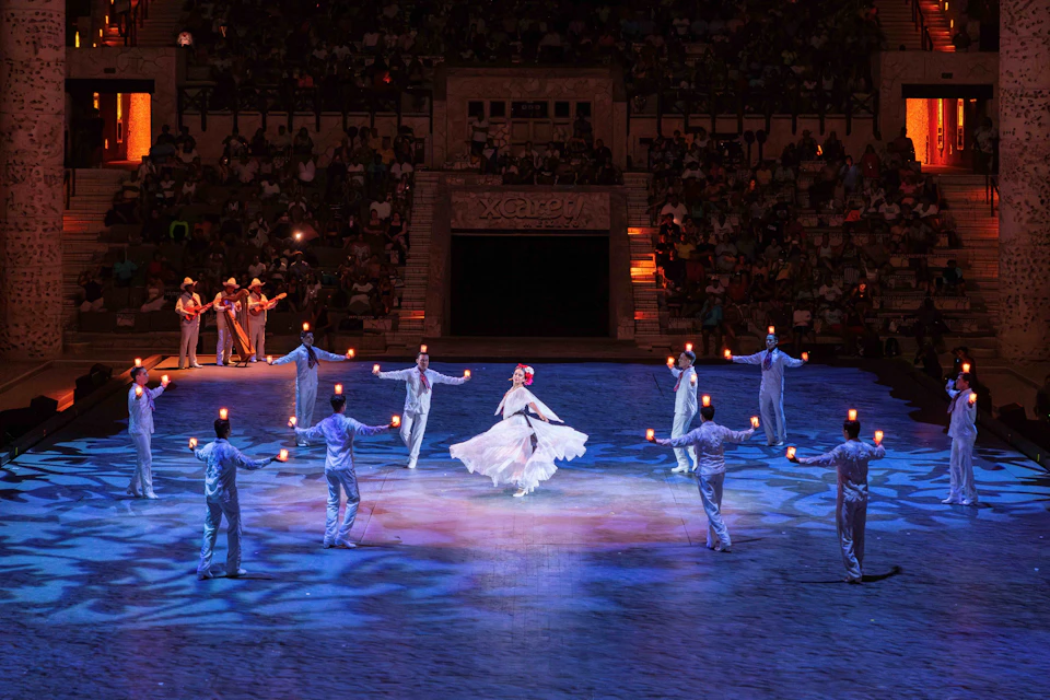 Performers in traditional attire dance in a well-lit arena with a large audience watching in the background.