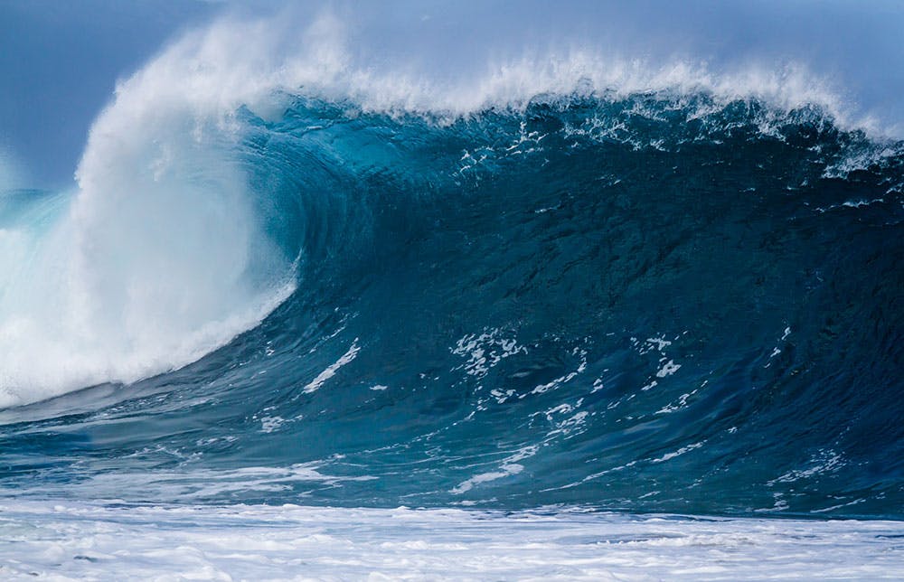 Large ocean wave curling, with white foam at the crest and base, under a cloudy sky.