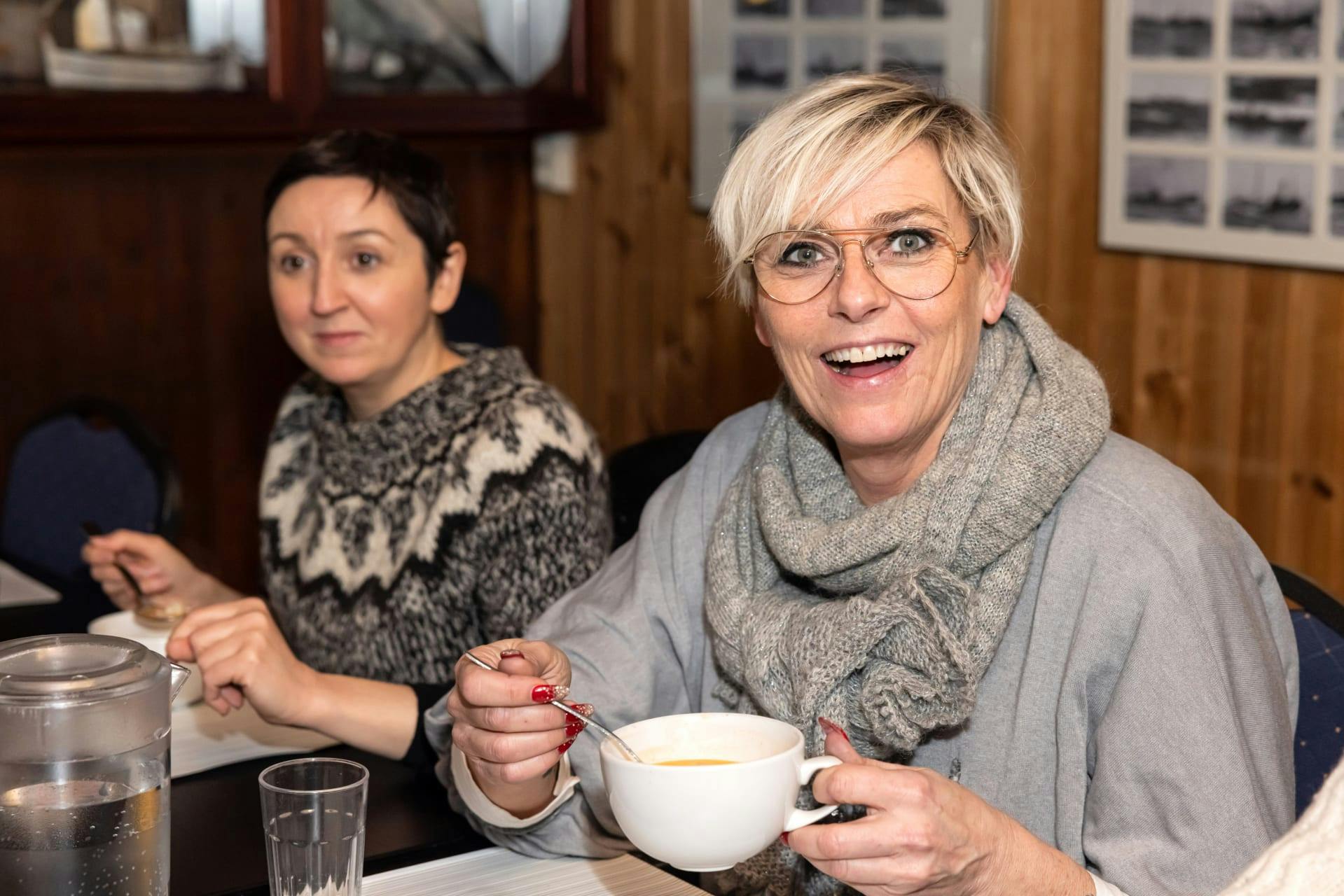 Two women sitting at a table, one smiling and holding a cup of soup, the other wearing a fair isle sweater.