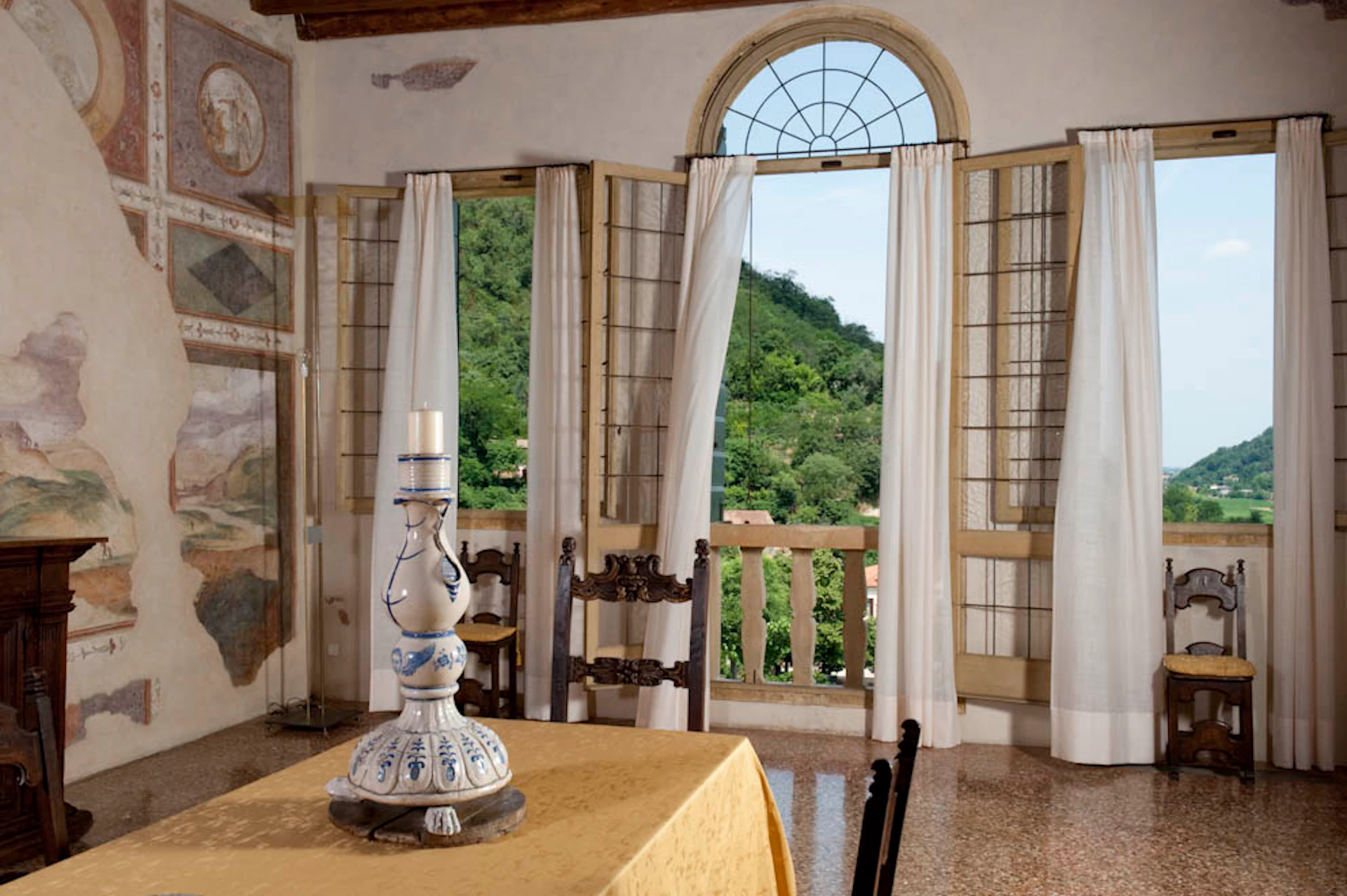 Room with large arched windows, open shutters, a table with a candle holder, and a view of a lush green landscape outside.