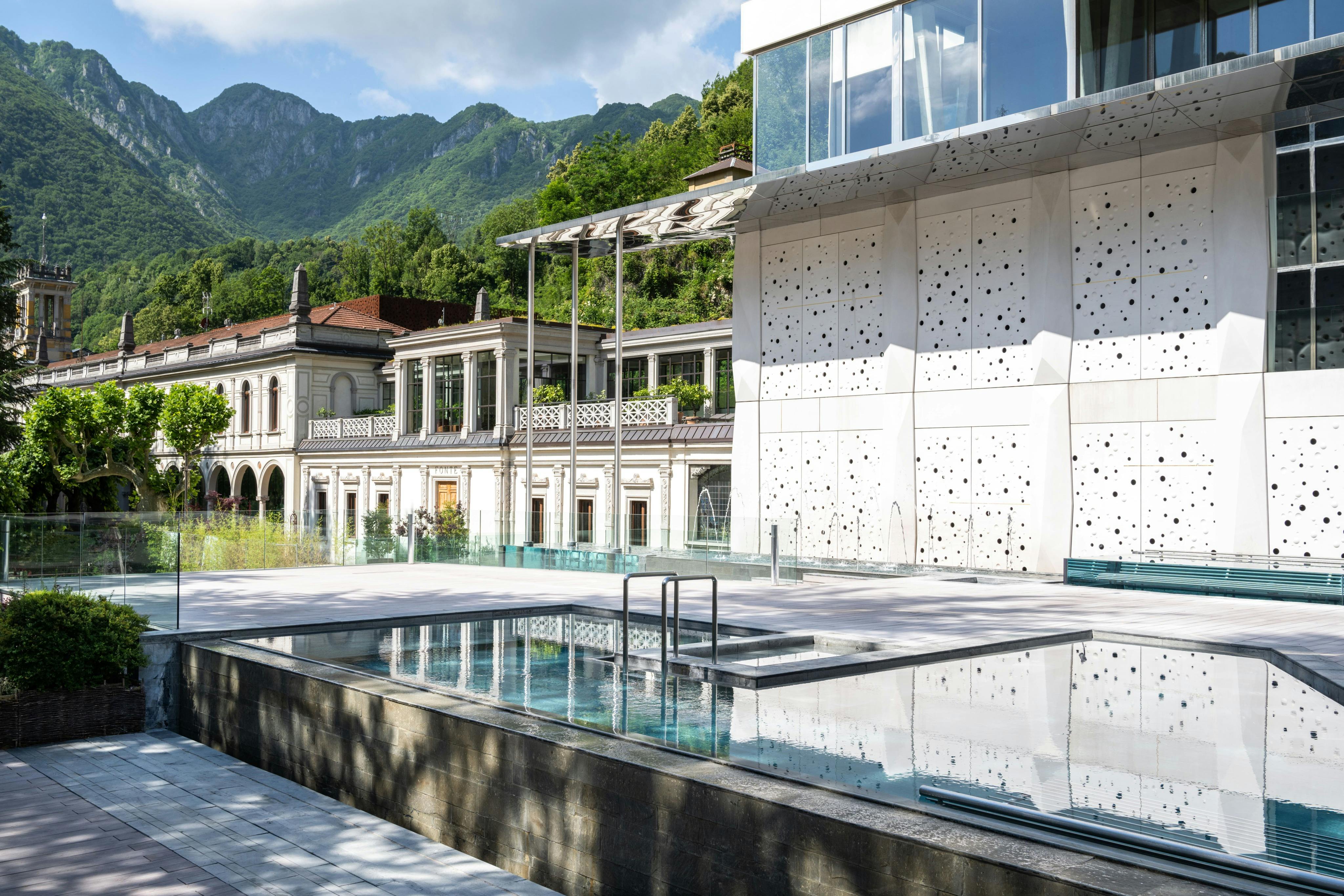 Edificio moderno con piscina in primo piano, circondato da verde e montagne sotto un cielo parzialmente nuvoloso.