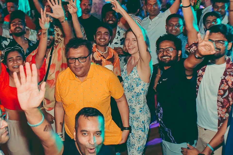 A crowd of people at an outdoor nighttime party, smiling and raising their hands, with city lights in the background.