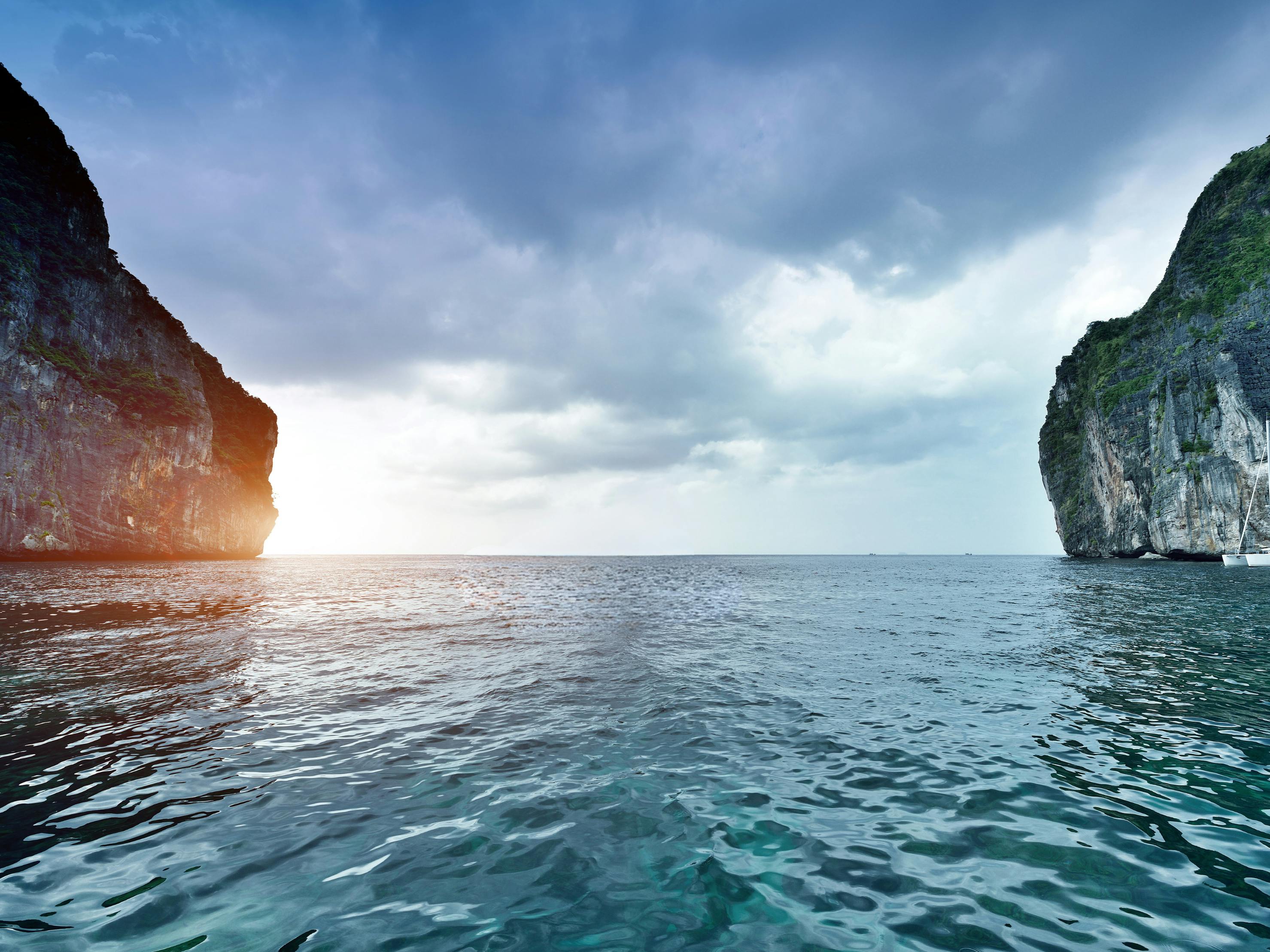 Mer calme avec des falaises imposantes de part et d'autre sous un ciel nuageux, la lumière du soleil apparaissant à l'horizon.