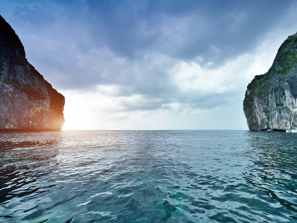 Calm sea with towering cliffs on either side under a cloudy sky, sunlight peeking through on the horizon.