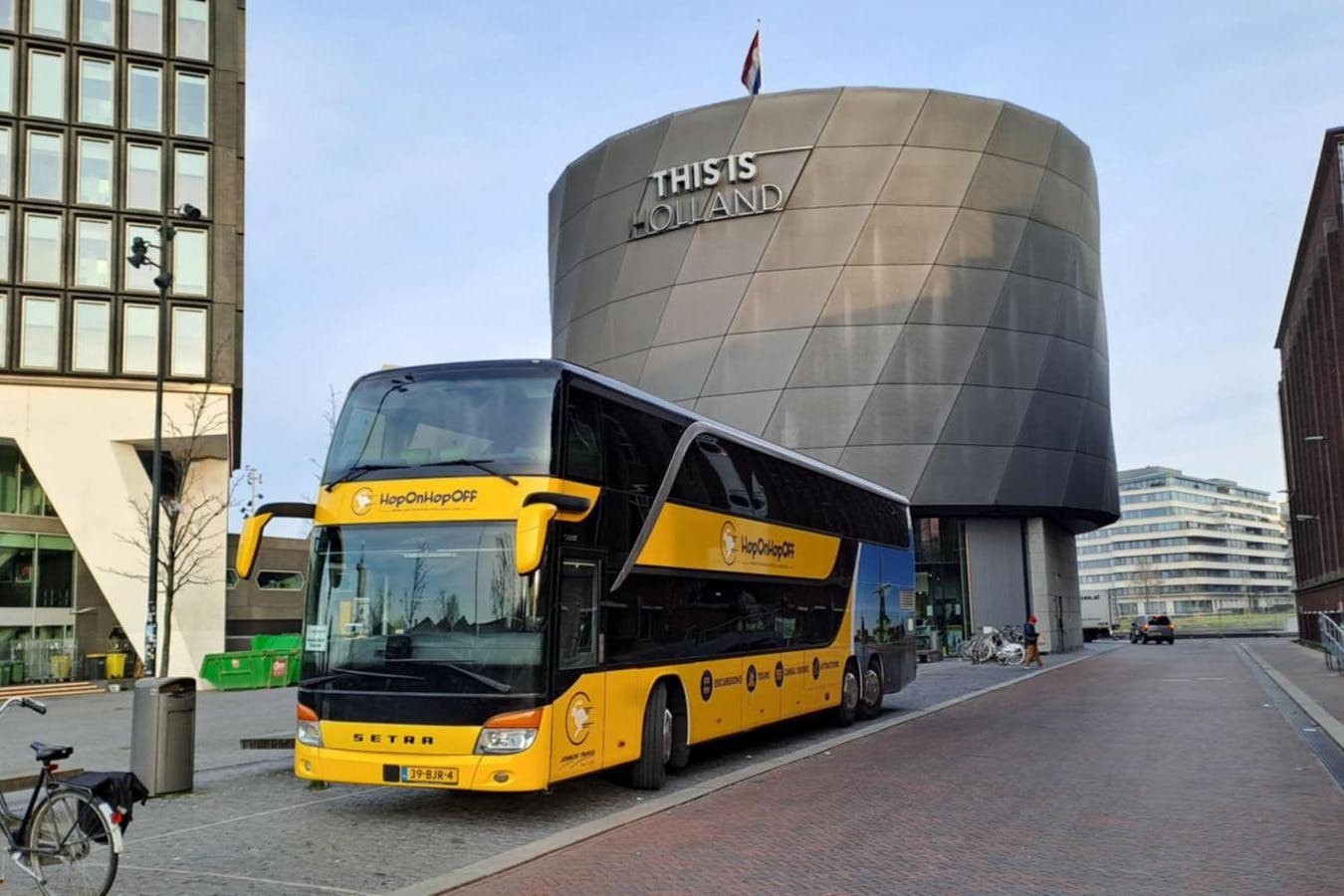 A yellow double-decker "Hop On Hop Off" tour bus parked in front of a modern building with "THIS IS HOLLAND" written on it.