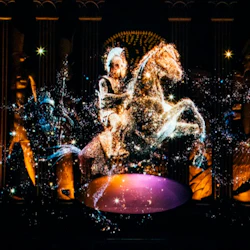 En glödande holografisk figur av en ryttare på en häst, omgiven av glitter, framför en bakgrund av statyer och pelare.