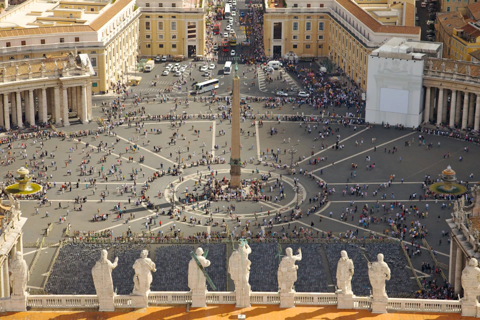 A large plaza with an obelisk at the center, surrounded by crowds, seen from above with statues in the foreground.