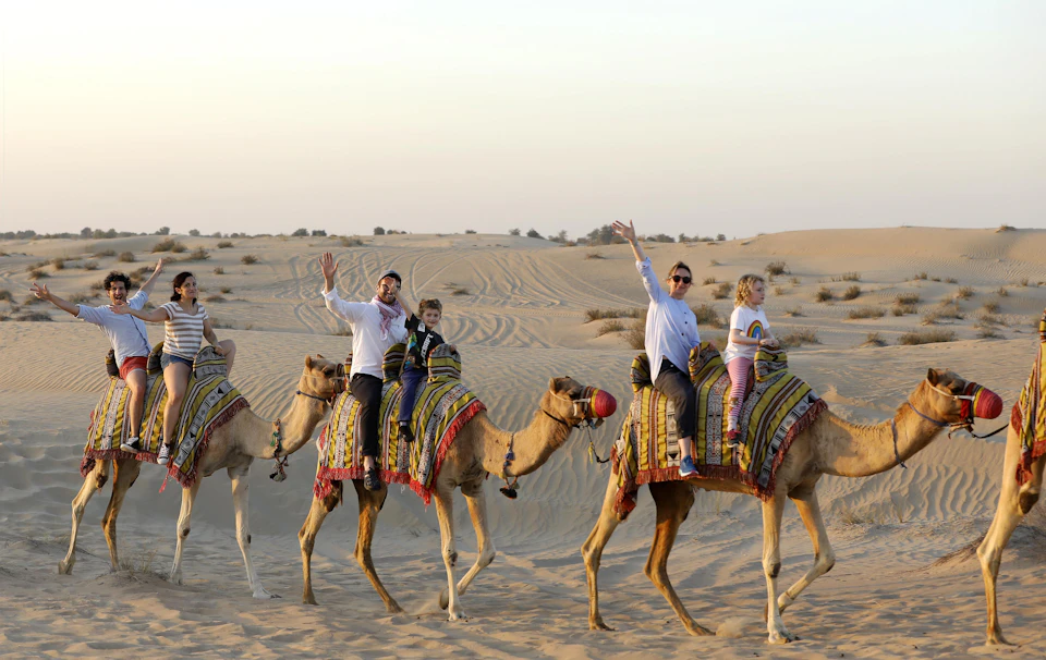 People riding camels adorned with decorative blankets in a desert, smiling and waving against a backdrop of sand dunes.