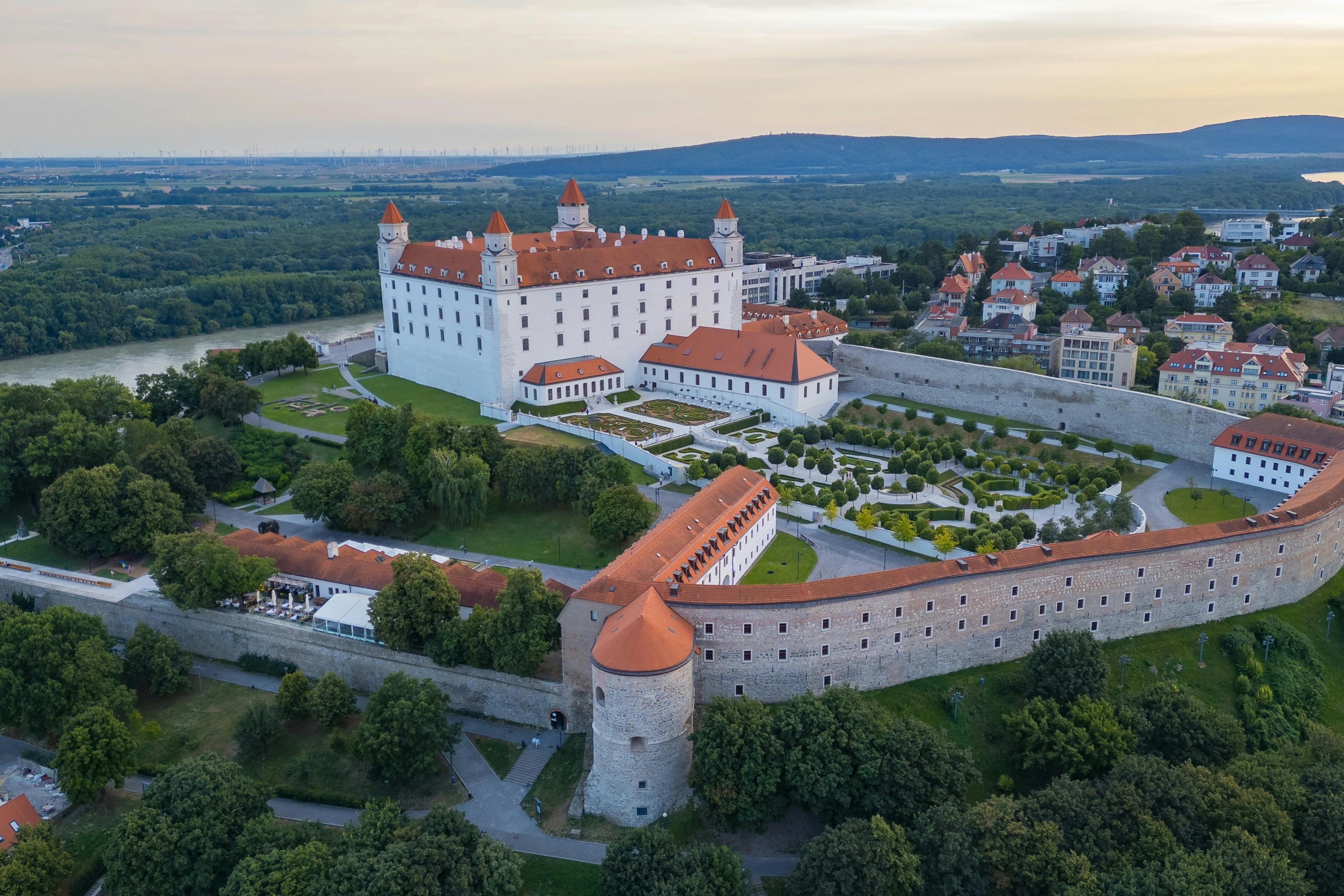 An aerial view of a historic castle with red roofs, surrounded by gardens and greenery, in a hilly urban landscape.