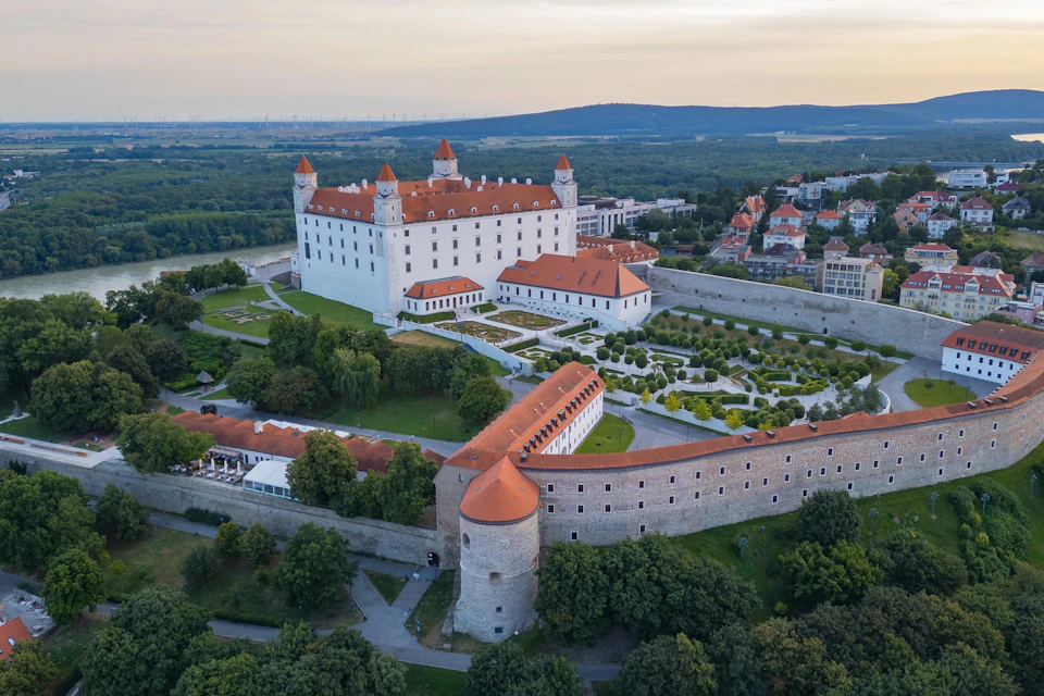 An aerial view of a historic castle with red roofs, surrounded by gardens and greenery, in a hilly urban landscape.