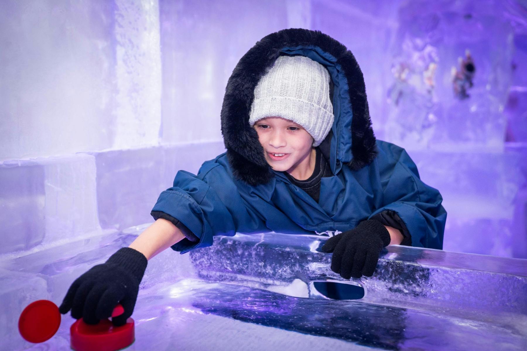 파란색 파카와 회색 비니를 입은 어린이가 얼음을 주제로 한 환경에서 보라색 조명으로 빛나는 얼음 블록을 가지고 놀고 있습니다.