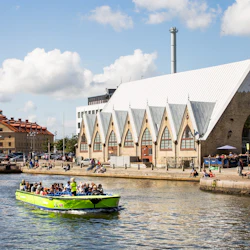 En grön turbåt med människor passerar framför en byggnad vid vattnet med en triangulär takdesign på en solig dag.