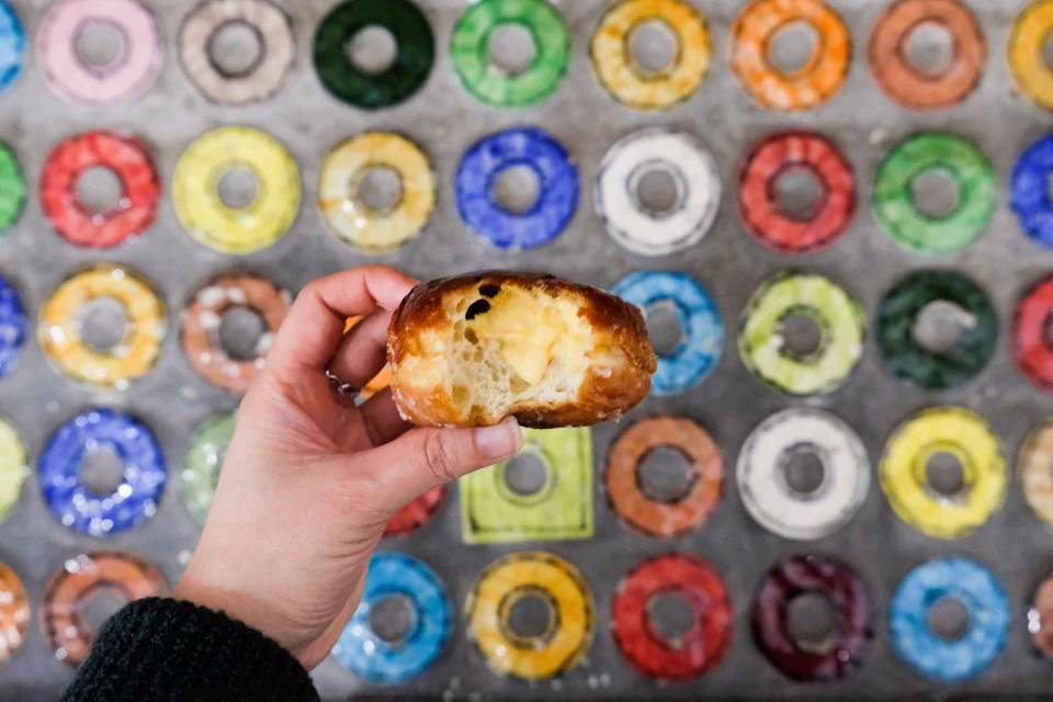 Hand holding a bitten glazed doughnut, in front of a background of various colorful doughnuts arranged in rows.