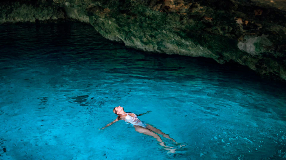 Person floating in clear blue water with rocky cave ceiling above.