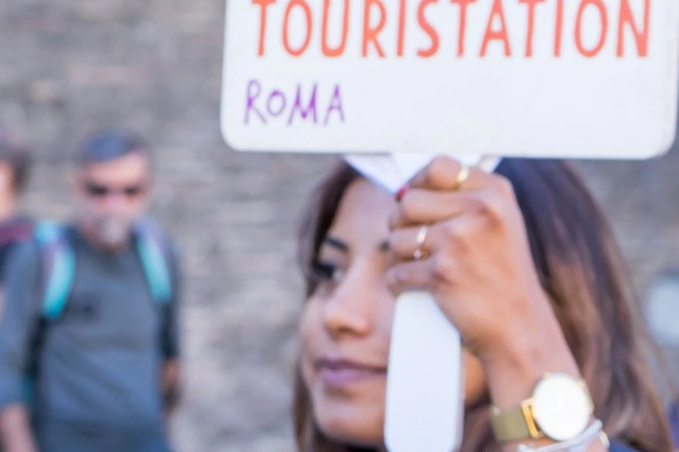 A tour guide in a navy blazer holds a "Touristation Roma" sign, leading a group of attentive tourists against a historic backdrop.