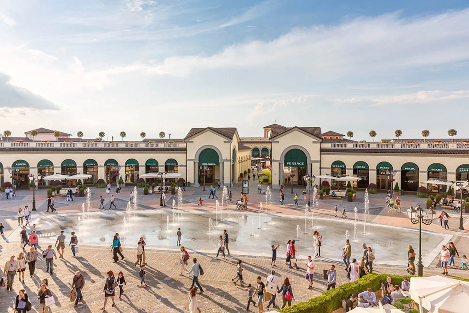 People walking around a plaza with water fountains and shops, including a Versace store, under a clear, blue sky.