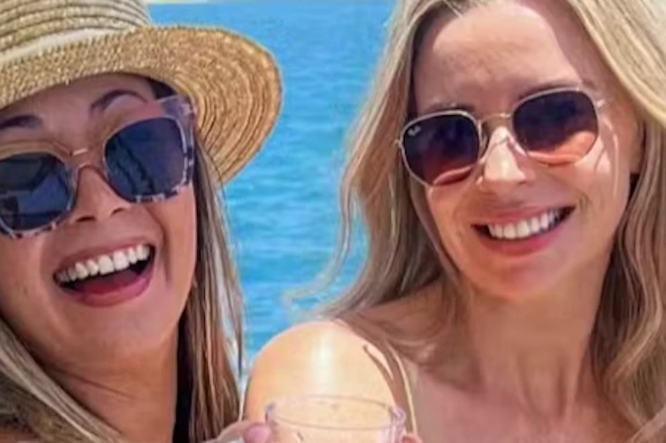 Three women smiling and holding drinks on a boat with a body of water and urban skyline in the background.
