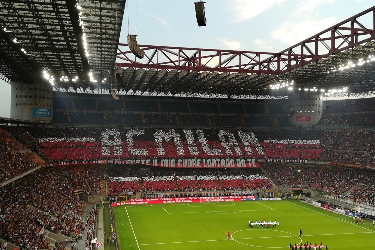 A soccer stadium with fans holding cards forming the text "AC MILAN" in red, black, and white, with players on the field.