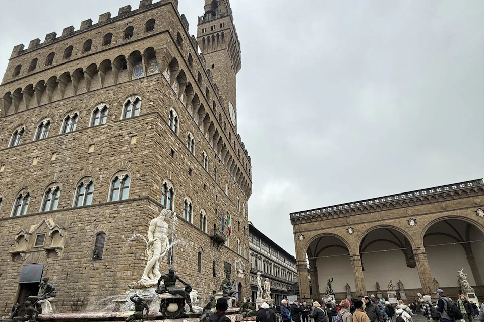 A historical plaza with a stone building, statues, and people walking around under a cloudy sky.