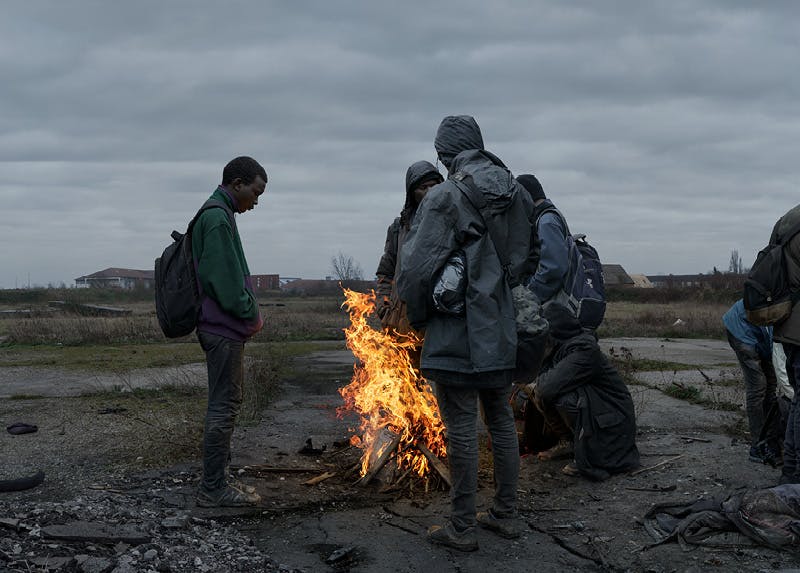 Several people wearing jackets and backpacks stand around a small fire in a desolate outdoor area under a cloudy sky.