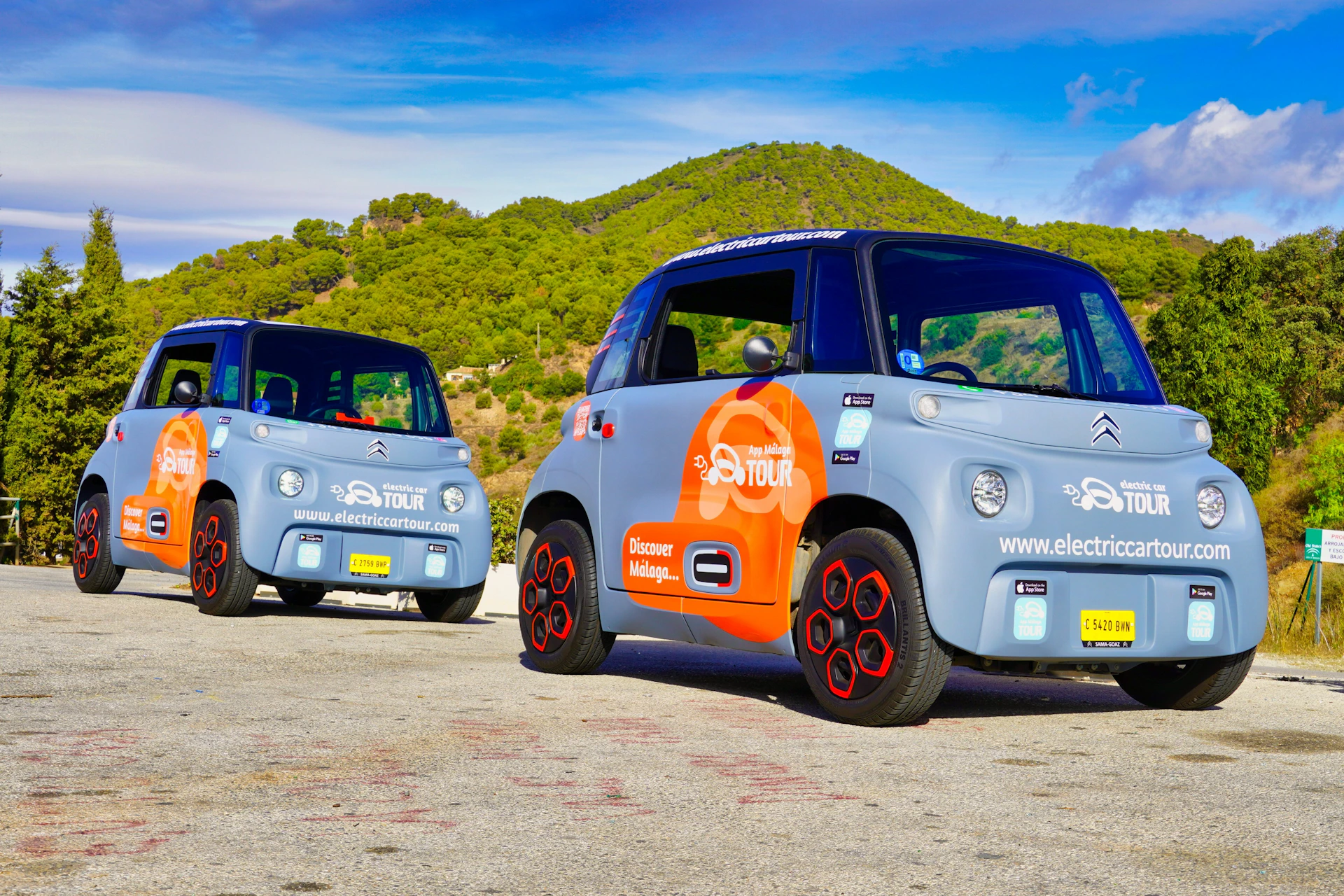 Two small electric cars with colorful branding parked on a road, with trees and hills in the background.
